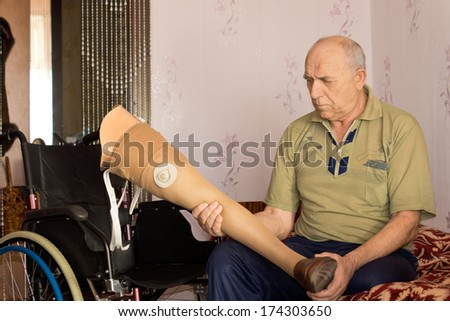 Senior disabled man sitting on a bed alongside his wheelchair holding a prosthetic leg or artificial limb