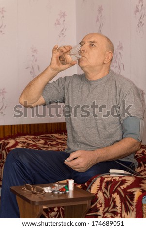Senior man swallowing down his medication as he sits at a small table with an assortment of tablets and pills