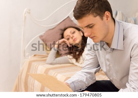 Profile close up view of a handsome young man reading a letter in the bedroom sitting on the edge of the bed watched by a young woman lying on the bedclothes