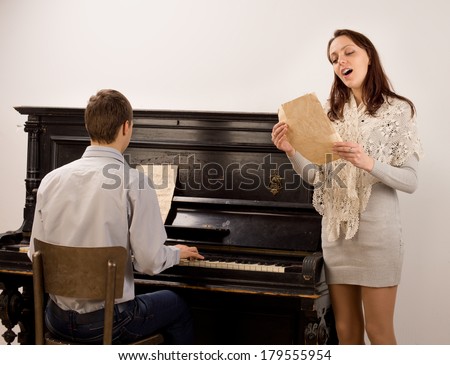 Young woman singing a solo song standing alongside a young man playing an upright piano singing with obvious enjoyment