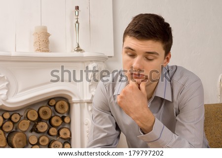 Sad depressed young man in a stylish shirt sitting in front of an ornate marble fireplace with downcast eyes and his chin on his hand