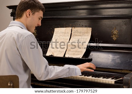 Young man having fun playing the piano smiling as he plays a melody from an old music score on a wooden upright piano