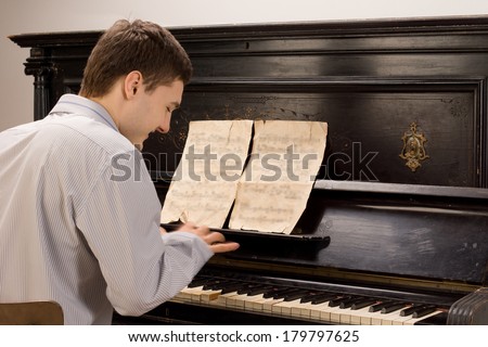 View from the rear of a young man smiling as he plays the piano using an old vintage music score on an upright wooden piano