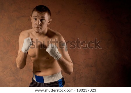 Pugnacious determined young boxer standing with his fists at the ready in front of his chest facing towards the camera on a graduated brown background with copyspace