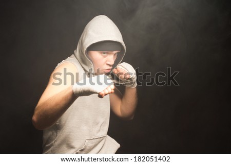 Pugnacious young boxer wearing a hooded top raising his bandaged fists in a fight in a dark smoky atmosphere with copyspace