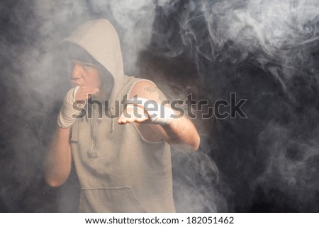 Young boxer working out in a smoky room in a hooded grey top standing with his bandaged hands raised waiting and watching