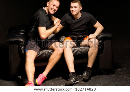 Two happy fit young men having a friendly arm wrestle as they sit together on a black leather couch in the darkness