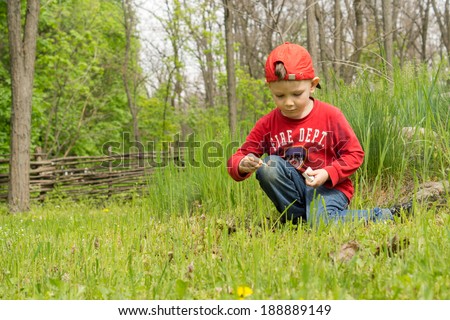 Little boy dressed in red fire department sweater kneeling down in the green grass lighting his own small campfire, with copyspace over woodland