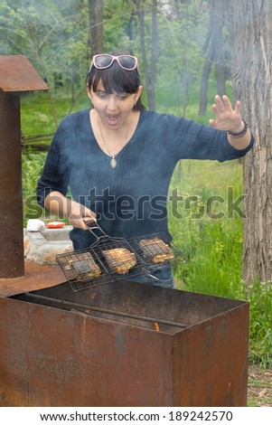 Woman cooking on a BBQ calling out as the flames start to flare up from the fat dripping from the meat as she lifts the grill clear of the heat