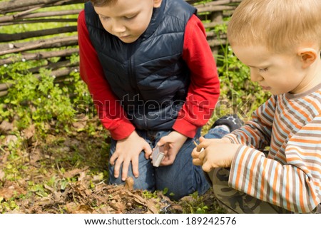 Two small boys trying to light a fire kneeling down on the ground over a pile of twigs and leaves with a box of matches