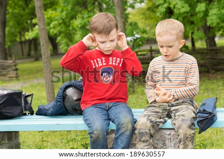 Upset little boy covering his ears as his cute little friend tries to console him as they sit together waiting on a rustic wooden bench with their luggage