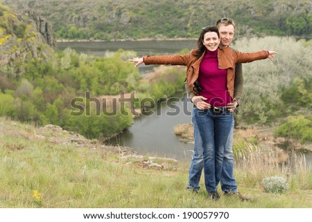 Happy romantic young couple celebrating an enjoyable day out in the countryside with the man hugging the woman from behind as she smiles with outstretched arms overlooking a scenic valley and river