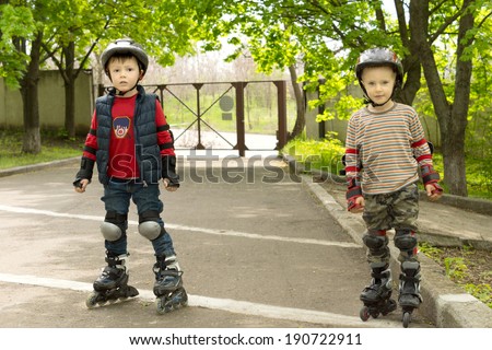 Two small boys kitted out for roller skating wearing roller blades, knee pads and safety helmets as they stand on a road in front of a park entrance