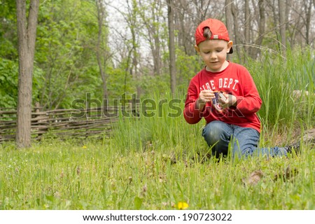 Handsome little boy trying to light a campfire kneeling down in the green grass in woodland in front of a pile of twigs and wood striking a match