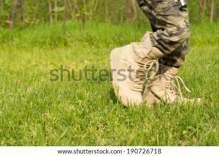 Little boy wearing camouflage pants in borrowed oversized lace-up boots walking across green grass, close up of his feet