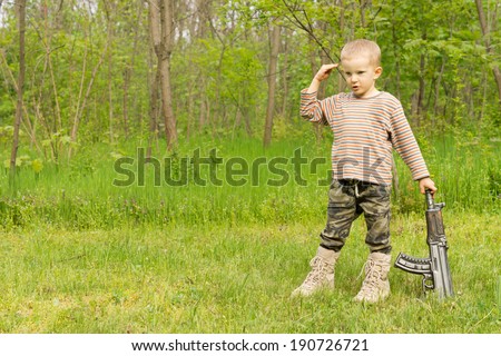 Little boy playing at soldiers standing saluting in camouflage pants and boots trailing an automatic rifle at his side in a lush green country field with copyspace