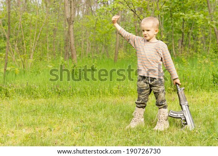 Macho little boy playing in camouflage pants and boots with an automatic rifle standing in a rural field raising his fist as he pumps the air