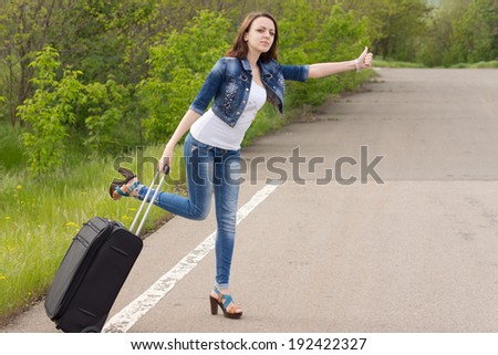 Desperate sexy beautiful young woman trying to thumb a lift holding her suitcase in one hand while leaning out into the road trying to attract the attention of passing motorists