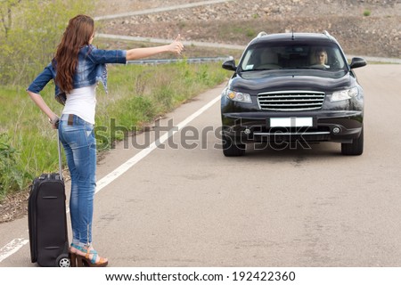 Trendy woman hitchhiking on a country road with her suitcase after breaking down at the roadside stepping into the road gesturing to a passing car