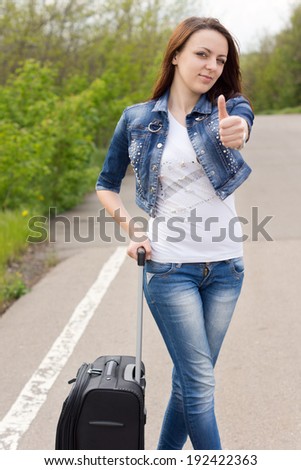 Smiling attractive young woman giving a thumbs up as she stands at the side of a rural road with her suitcase trying to thumb a lift