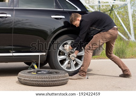 Young man struggling to change his car tyre as he battles with a wheel spanner to loosen the nuts on the hub standing putting his weight behind the effort