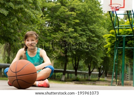 Beautiful confident young female basketball player sitting cross-legged on the outdoor court surrounded by leafy green trees with the ball waiting for play to commence