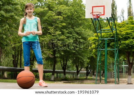 Young girl waiting to play a game of basketball standing on an outdoor asphalt court with the ball against a backdrop of green trees