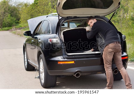 Man broken down at the roadside with his car searching in the boot for his tools and looking back over his shoulder at the camera