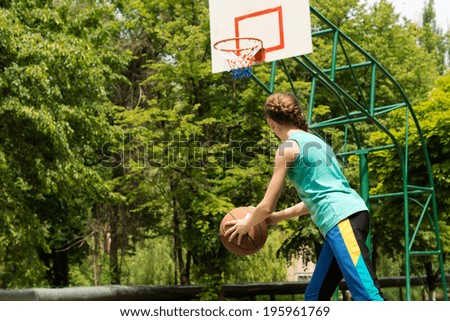 Sporty slender teenage girl playing basketball holding the ball in her hands as she moves forwards eyeing the goalpost on an outdoor court against green trees