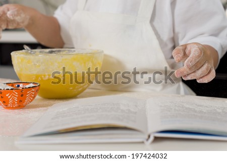 Young child baking a cake in the kitchen pointing with flour covered hands to the recipe book, close up of his hands, the book and a mixing bowl of ingredients