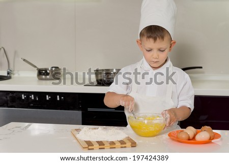 Little boy standing baking cakes in a toque and apron concentrating on mixing eggs and flour in a mixing bowl, with copyspace