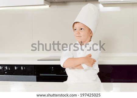 Confident smiling young boy in a chefs uniform standing in the kitchen in front of the stove with his arms folded