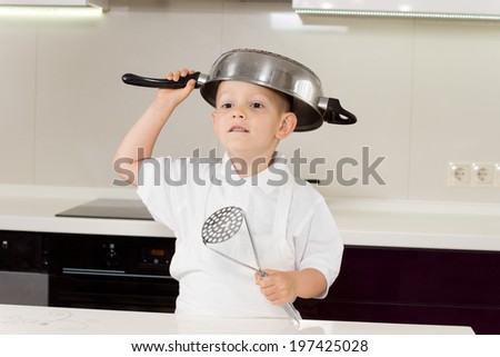 Little boy in a white apron clowning around with kitchen utensils standing with a saucepan upended on his head and a ladle in his hand