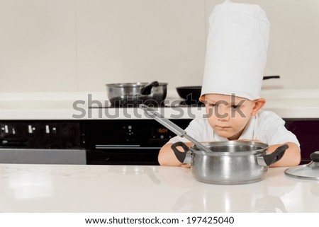Little boy cooking in his apron and chefs hat bending down to smell the food in the saucepan with a thoughtful expression