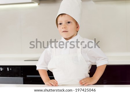 Proud confident little boy in a chefs uniform standing in his white apron and toque with his hands on his hips and a serious expression