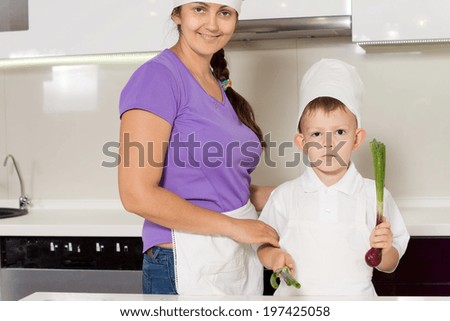 Smiling mother and son in chefs outfits standing together in the kitchen holding fresh onions as she teaches him the basics of cooking