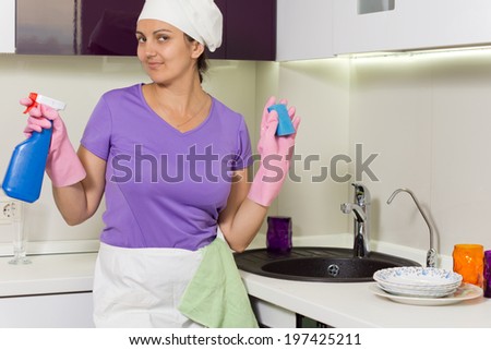 Smiling housewife holding up detergent and soap smiling playfully at the camera as she stands at the sink in her apron and cap