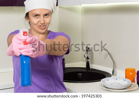 Playful housewife in a white cap and rubber gloves standing at the sink aiming the spray bottle at the camera