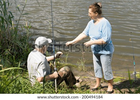 Elderly couple fishing on a freshwater lake with the man seated on the bank with his rod as his wife assists him