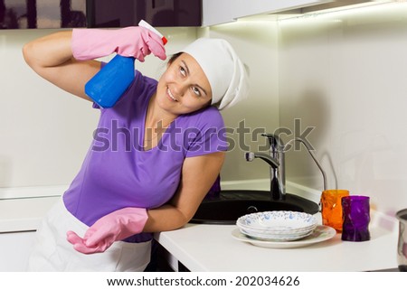 Silly woman holding spray cleaner to her head near the kitchen sink