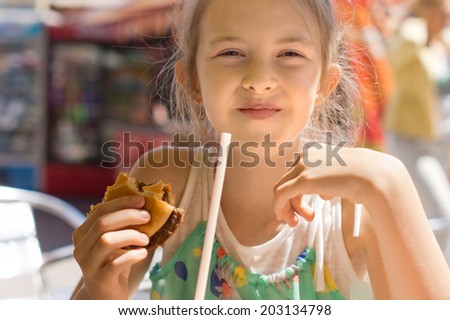 Happy cute girl eating a tasty hamburger with ground beef meat while having the lunch at fast-food restaurant, in a summer sunny day