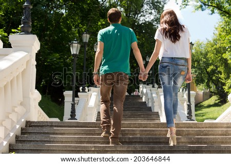 Affectionate young couple walking away from the camera up a flight of steps in a park shaded by trees holding hands