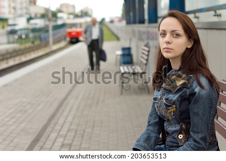Lonely beautiful girl, with a deep look and a sad facial expression, sitting on a bench while waiting in an urban railway station