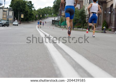 Low angle view from tarmac level of the legs of a group of road runners in a race running along an urban road past buildings and parked cars