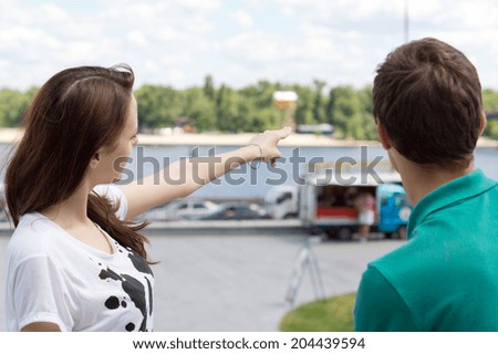 Attractive young woman pointing forward showing the right direction to a male tourist, outdoors, downtown, in front of a watercourse