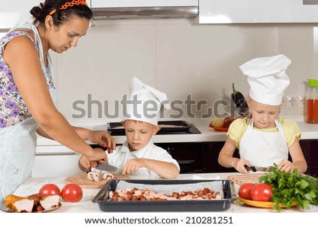 Mother teaching her children to cook homemade pizza guiding the little boy as he chops mushrooms with his sister