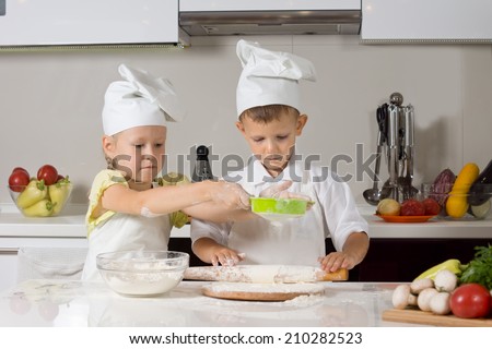 Cute small boy and girl wearing cooks uniforms baking together in the kitchen working a a team making and rolling out dough for pizza bases