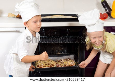 Two happy young children putting their homemade pizzas in the oven turning back to smile with pride and enjoyment in their white cooks uniforms