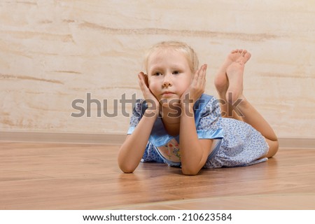 Bored little girl lying on the wooden floor with her bare feet up in the air and her chin resting on her hands as she stares ahead of her with a fed up expression