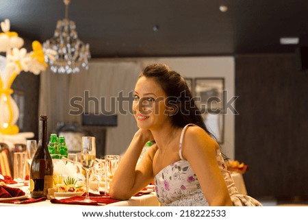Beautiful woman sitting alone at a formal dinner table set with elegant place settings, glassware and champagne leaning her chin on her hand giving the camera a warm friendly smile